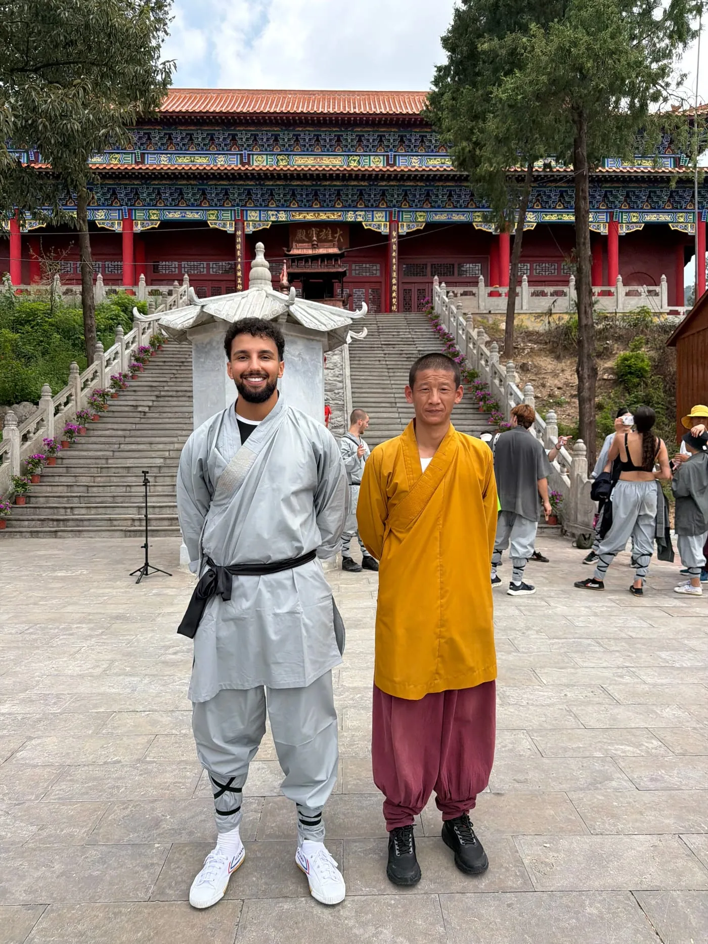 Yassine, en tenue de kung‑fu grise, pose aux côtés du maître en robe safran, au pied des escaliers d’un temple.
