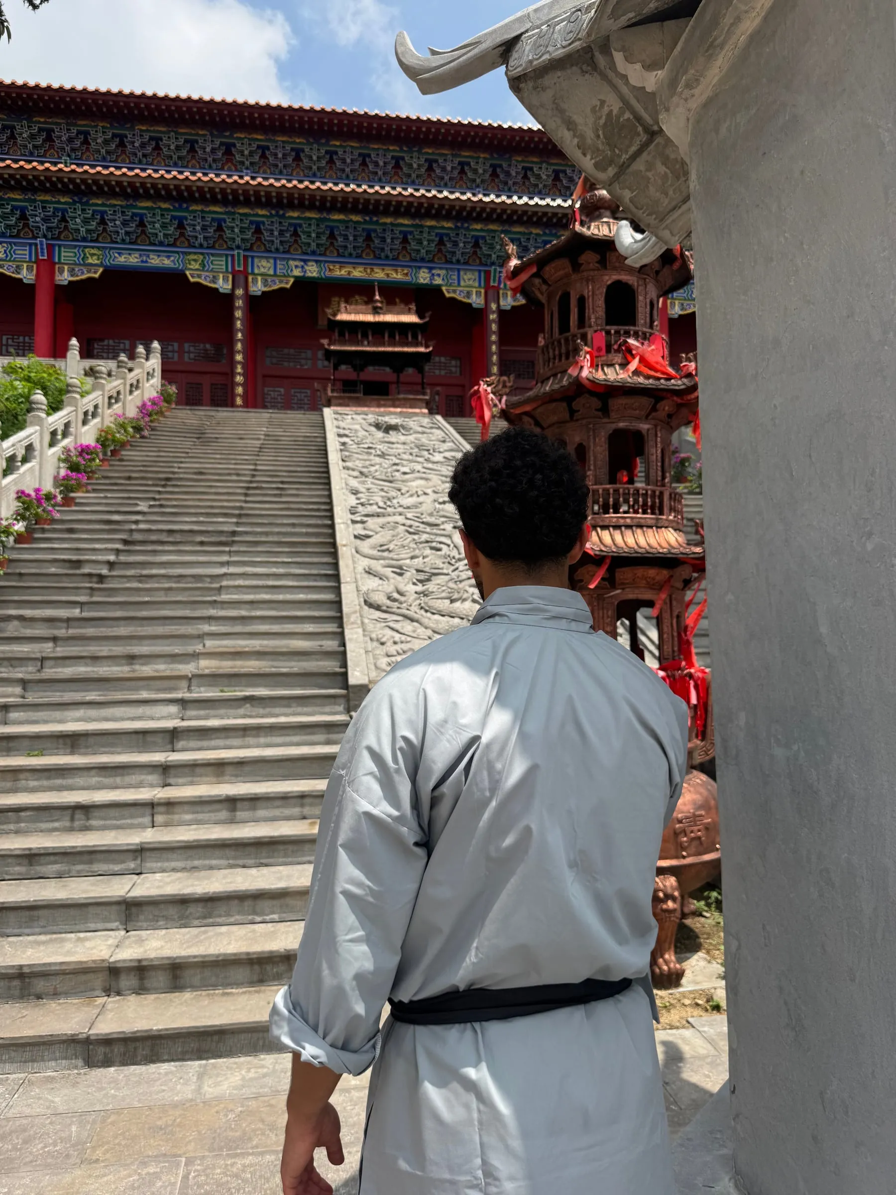 Yassine, de dos, en tenue de kung‑fu grise devant le grand escalier de pierre d’un temple aux colonnes rouges, près de Tengzhou.